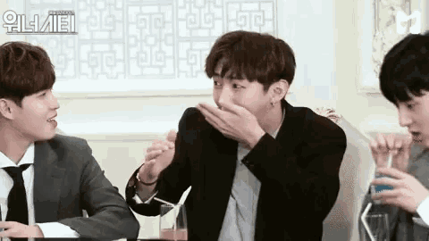 a group of young men in suits and ties are sitting at a table . one of the men is covering his mouth with his hand .