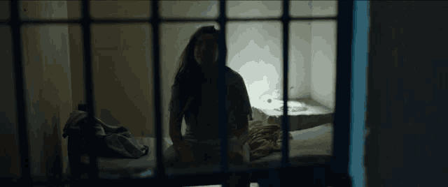 a woman is sitting on a bed in a dark room behind bars in a jail cell .