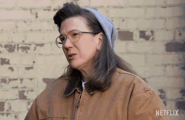 a woman wearing glasses and a beanie is standing in front of a brick wall with netflix written on the bottom right