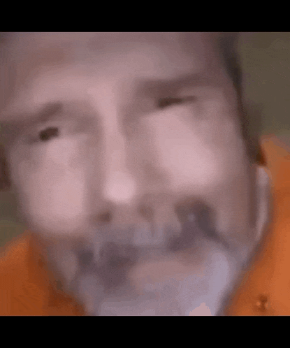 a close up of a man 's face with a beard wearing an orange shirt and looking at the camera .