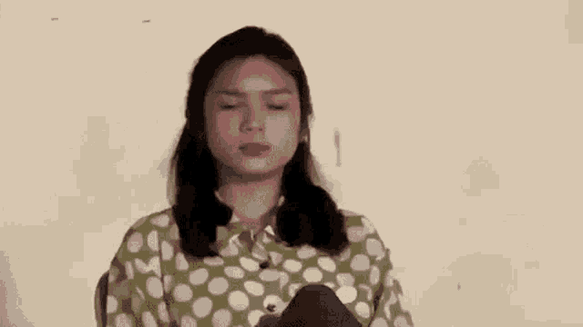a woman in a polka dot shirt is sitting down with her eyes closed and looking at the camera .