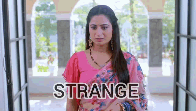a woman in a pink saree is standing in front of a window with the word strange written on the bottom .