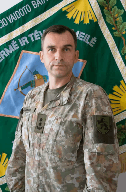 a man in a military uniform stands in front of a green banner that says " erme ir te "