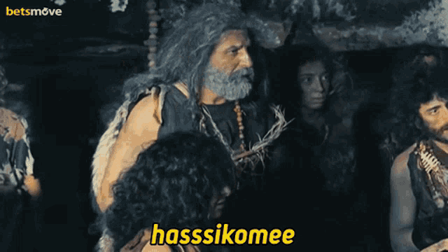 a man with long hair and a beard stands in front of a group of people and says " hasssikomee "
