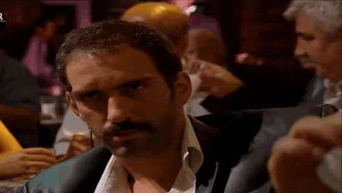 a man with a mustache is sitting at a table with a glass of wine in his hand .