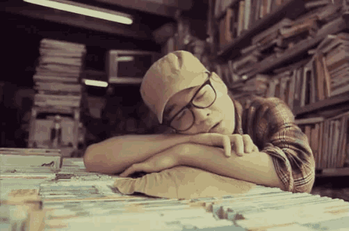 a man wearing glasses and a hat is sleeping on a pile of books in a library