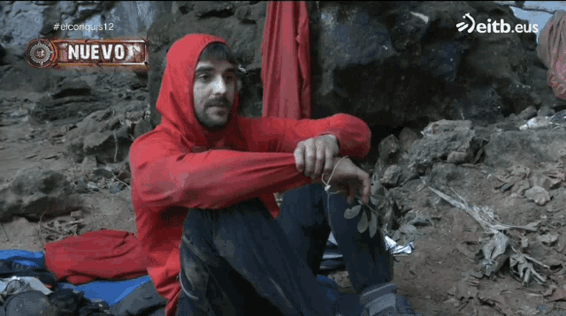 a man in a red hooded jacket sits on the ground with a sign that says nuevo