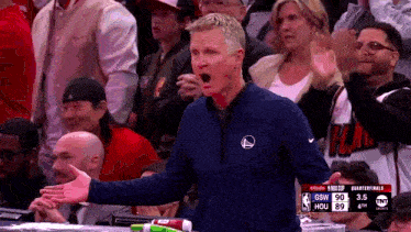 a man wearing a golden state warriors jacket stands in front of a crowd at a basketball game