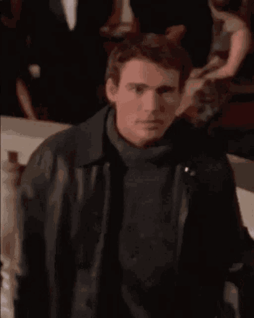 a man wearing a black leather jacket and a turtleneck is standing in front of a crowd of people .