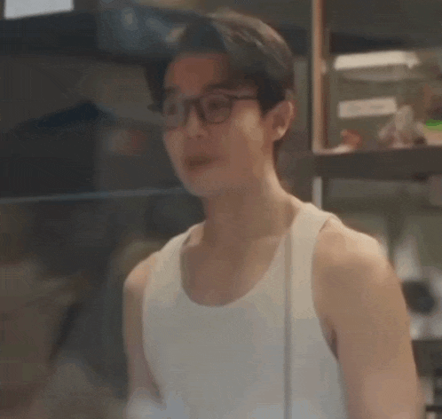 a young man wearing glasses and a white tank top is standing in front of a window .