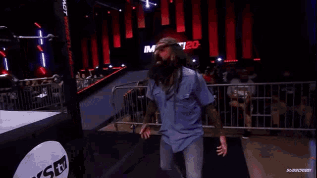 a man in a blue shirt stands in a wrestling ring with the word impact on the wall behind him