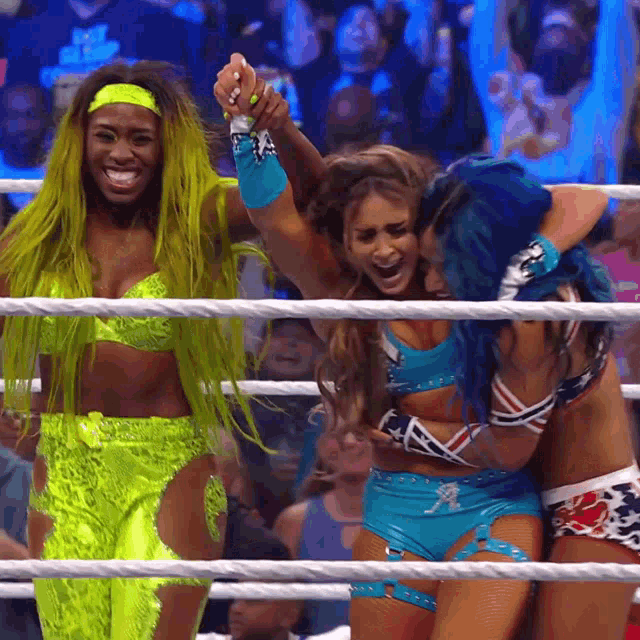 three women are hugging each other in a wrestling ring and one has blue hair