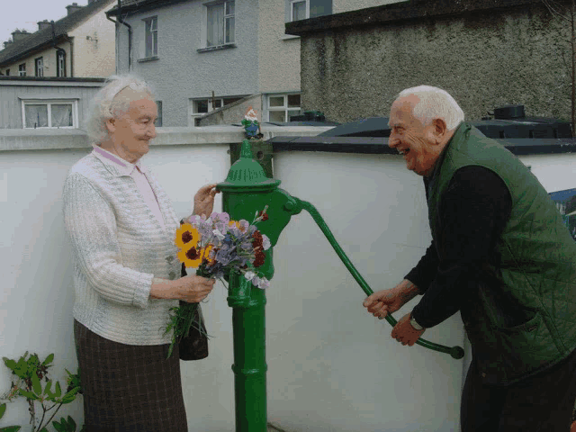 an elderly woman holds a bouquet of flowers while a man pumps water from a green pump