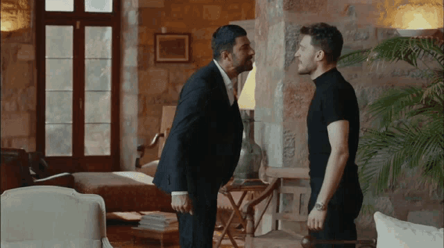 a man in a suit and tie stands next to another man in a black shirt in a living room