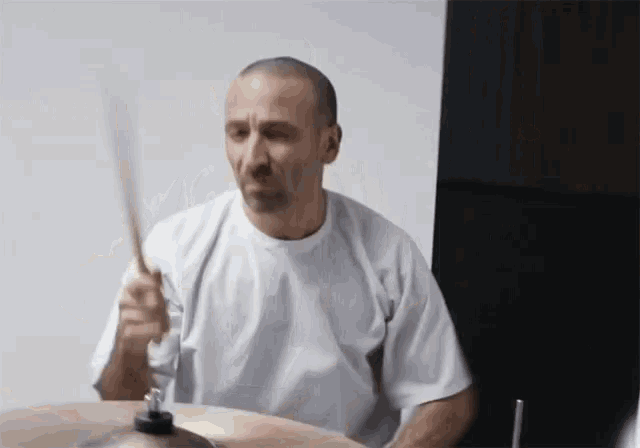 a bald man in a white shirt is playing a drum set