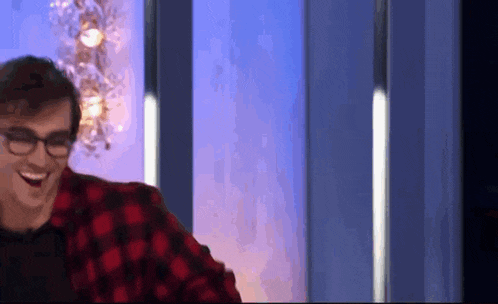 a man wearing glasses and a plaid shirt is laughing while sitting in front of a blue wall .