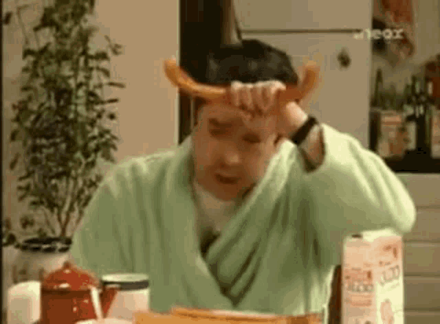 a man in a green robe is sitting at a table holding a hot dog on his head .