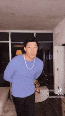 a man wearing a blue sweater and a gold chain is standing in a living room ..
