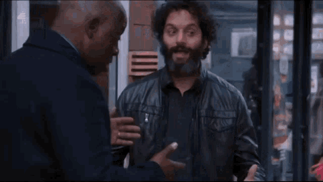 a man with a beard is talking to another man while wearing a leather jacket