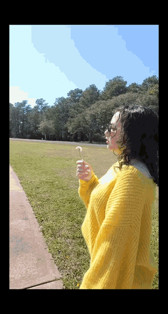 a woman in a yellow sweater blowing a dandelion in a field