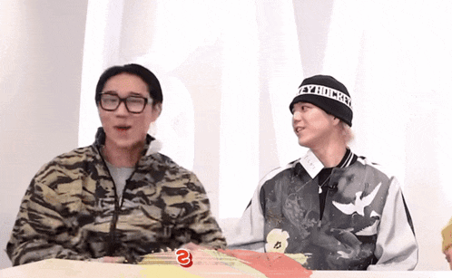 two men are sitting at a table and one has a beanie that says " hungry "