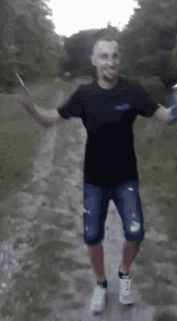 a man in a black shirt and blue shorts is walking down a dirt path holding a can of soda .