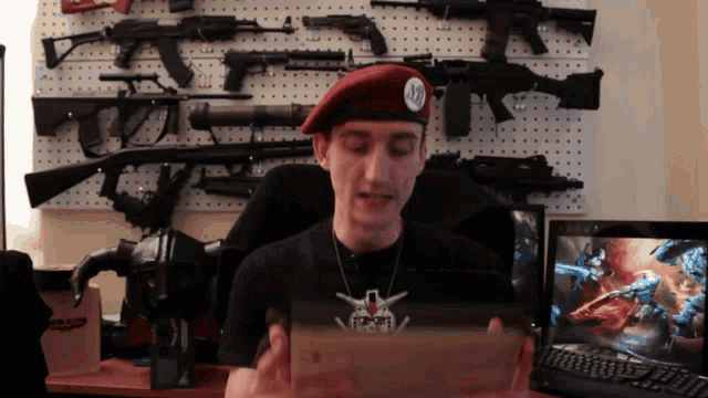 a man wearing a red beret holds a box in front of a wall filled with guns