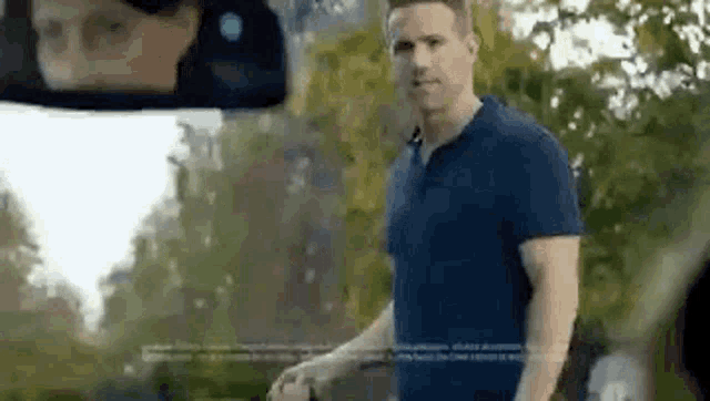 a man in a blue shirt is standing in front of a car and looking at the rear view mirror .