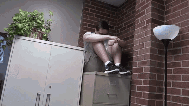 a man sits on a filing cabinet next to a brick wall