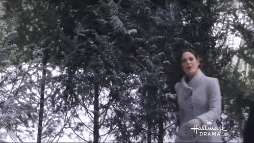 a woman in a grey coat is standing in front of a snow covered tree .