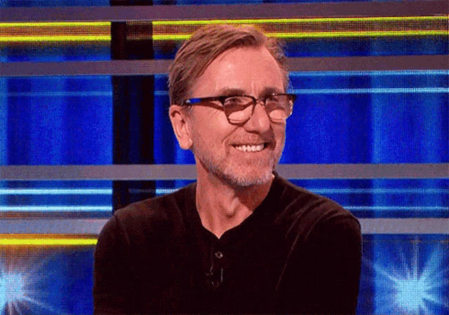 a man with glasses and a beard smiles in front of a blue background