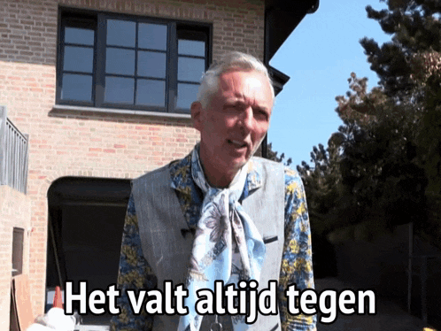 a man with a scarf around his neck is standing in front of a brick building with the words het valt altijd tegen above him