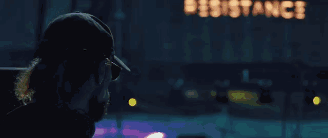 a man wearing a hat stands in front of a neon sign that says resistance