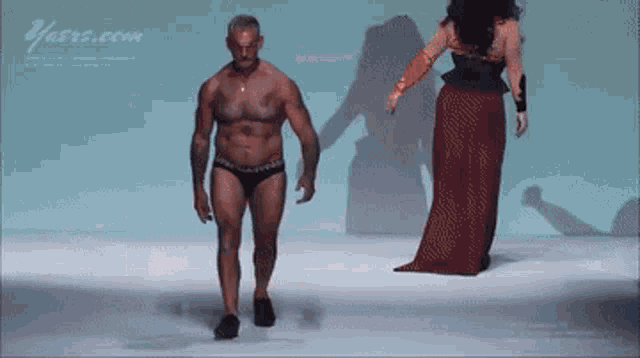 a man in underwear walks down a runway next to a woman in a long red dress