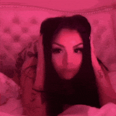a woman is laying on a bed with her hands on her head and a pink light behind her .