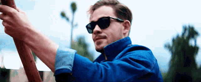 a man wearing sunglasses and a blue jacket is holding a wooden pole .