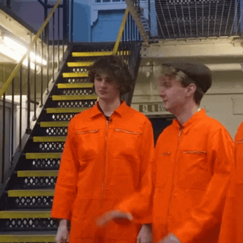 two men in orange jumpsuits are standing in front of a staircase