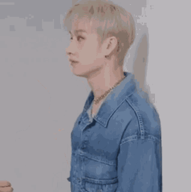 a young man wearing a denim jacket and a necklace is standing against a wall .