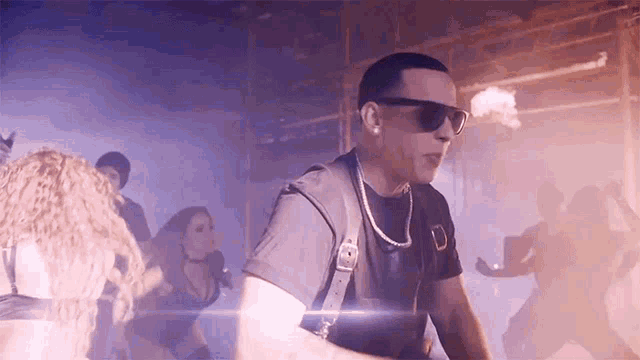 a man wearing sunglasses and a necklace is standing in front of a group of people dancing .