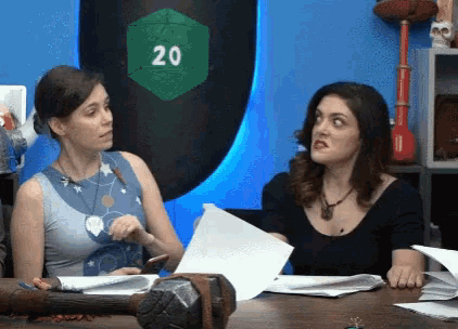two women are sitting at a table with papers and a green d20 on the wall behind them