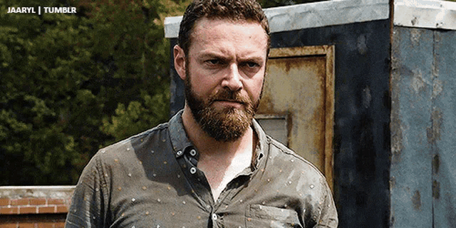 a man with a beard is standing in front of a brick wall and a rusty door with the words jaaryl tumblr below him