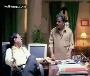 two men are sitting at a desk in front of a computer and talking .