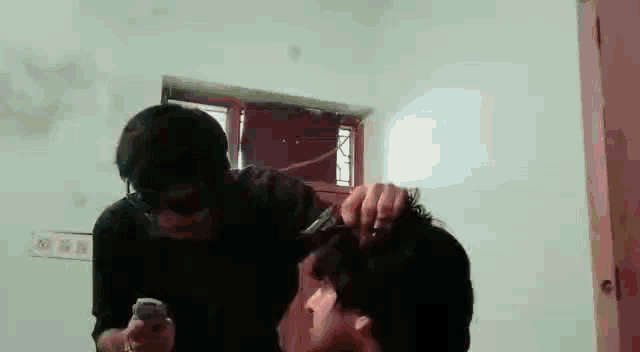 a man is cutting another man 's hair in a room while another man holds a cell phone .