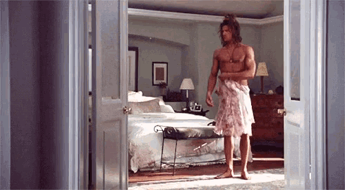 a shirtless man wrapped in a towel is standing in a bedroom .