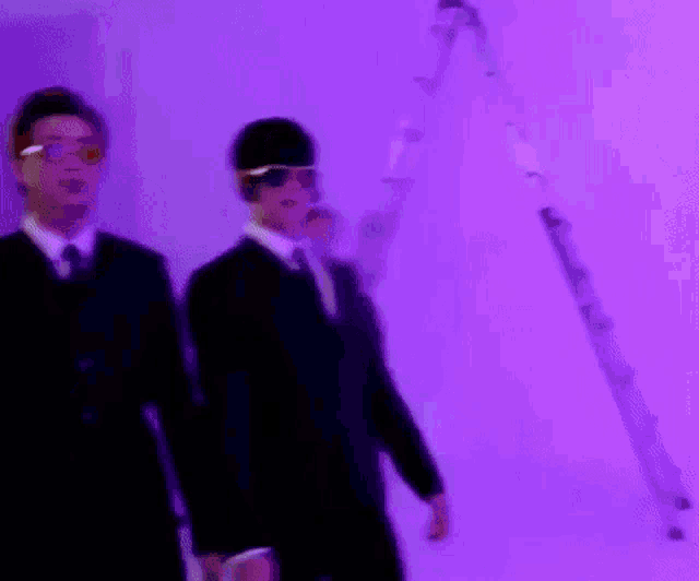 two men in suits and sunglasses are standing next to each other in front of a purple background .