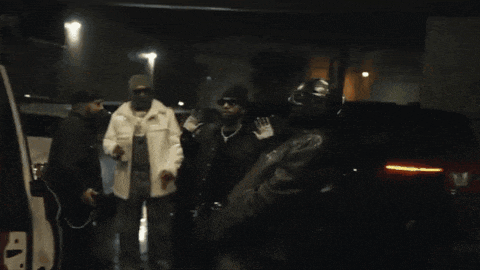 a group of men are standing in a dark room with a red car in the background