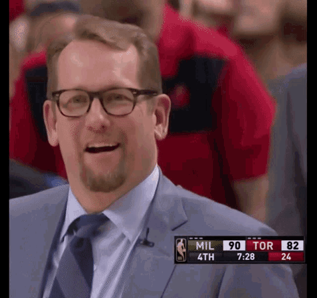 a man in a suit and tie smiles in front of a scoreboard that says mil 90 tor 32