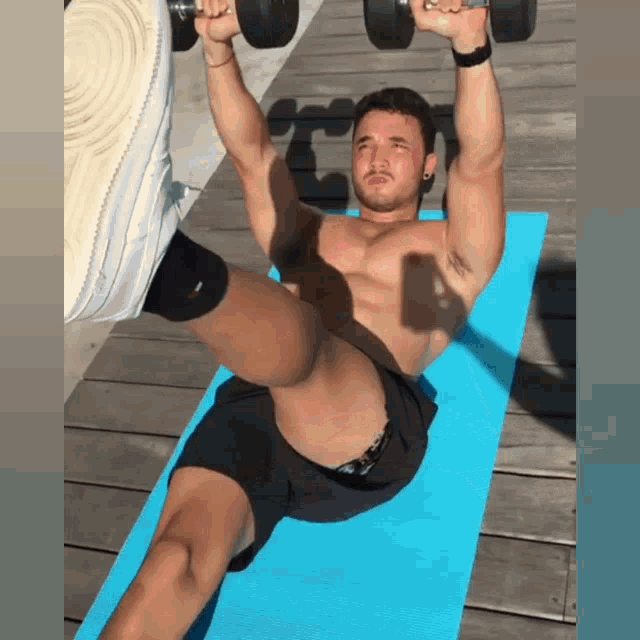 a shirtless man is lifting dumbbells over his head while laying on a blue mat