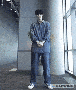 a man in a denim shirt and jeans is standing in front of a window with his hands on his hips .
