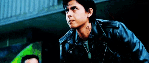 a young man wearing a black leather jacket and a hat is standing in front of a building .
